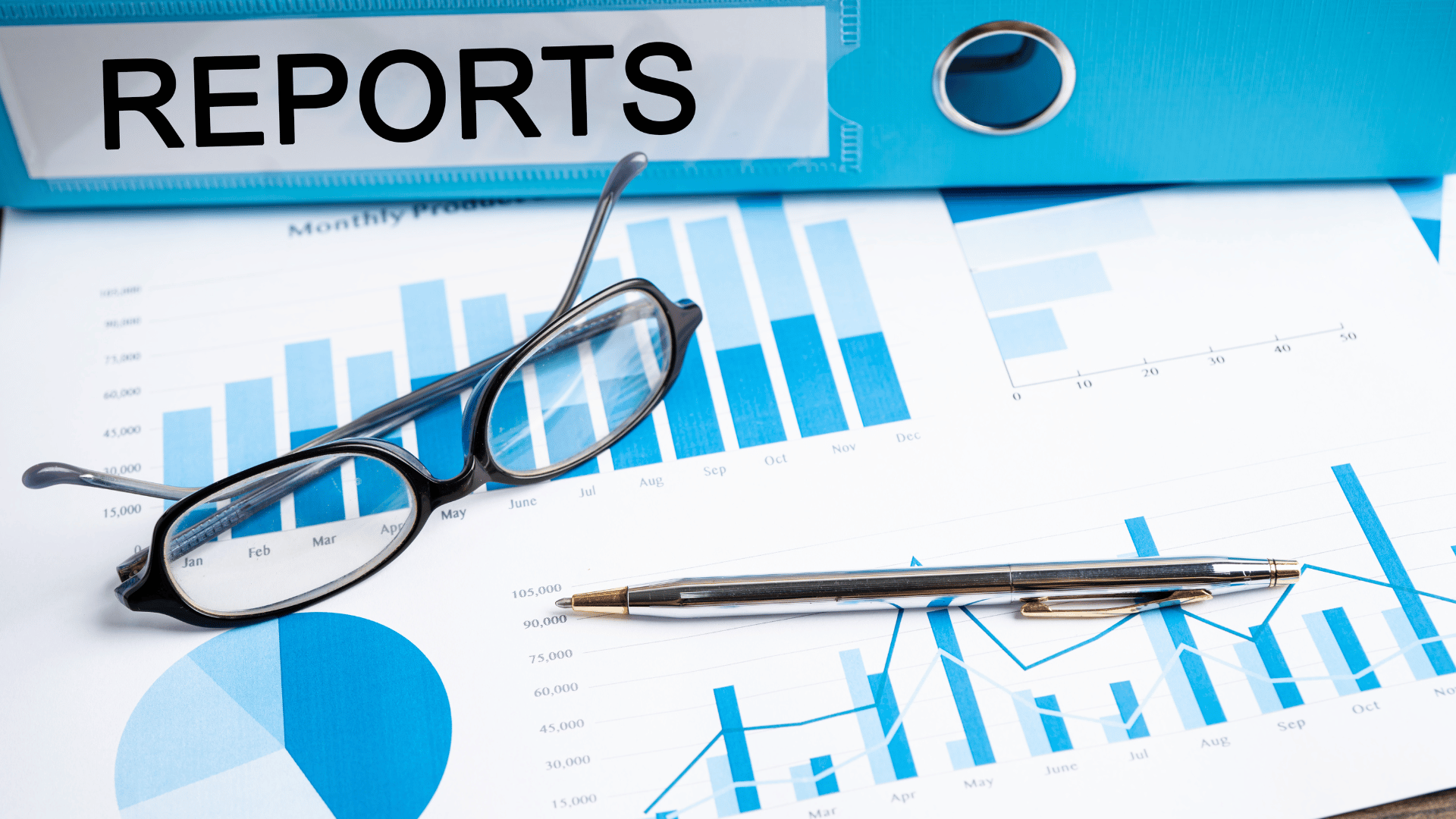 A blue binder sits on top of a few sheets of paper. The binder has white tape on it and says "report." Next to the papers, there's a pair of glasses and a pen. The sheets under the binder show diagrams of charts and reports.